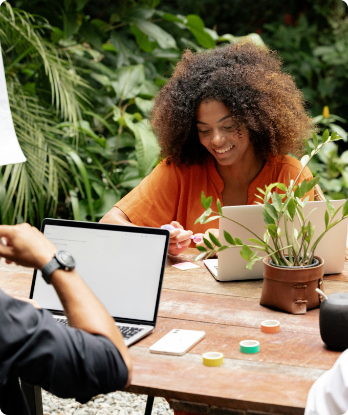 A smiling woman with curly hair in an orange shirt working on a laptop outdoors at a wooden table with plants and colored tape rolls.