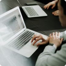 Person typing on a laptop keyboard at a table with another laptop and people in the background.