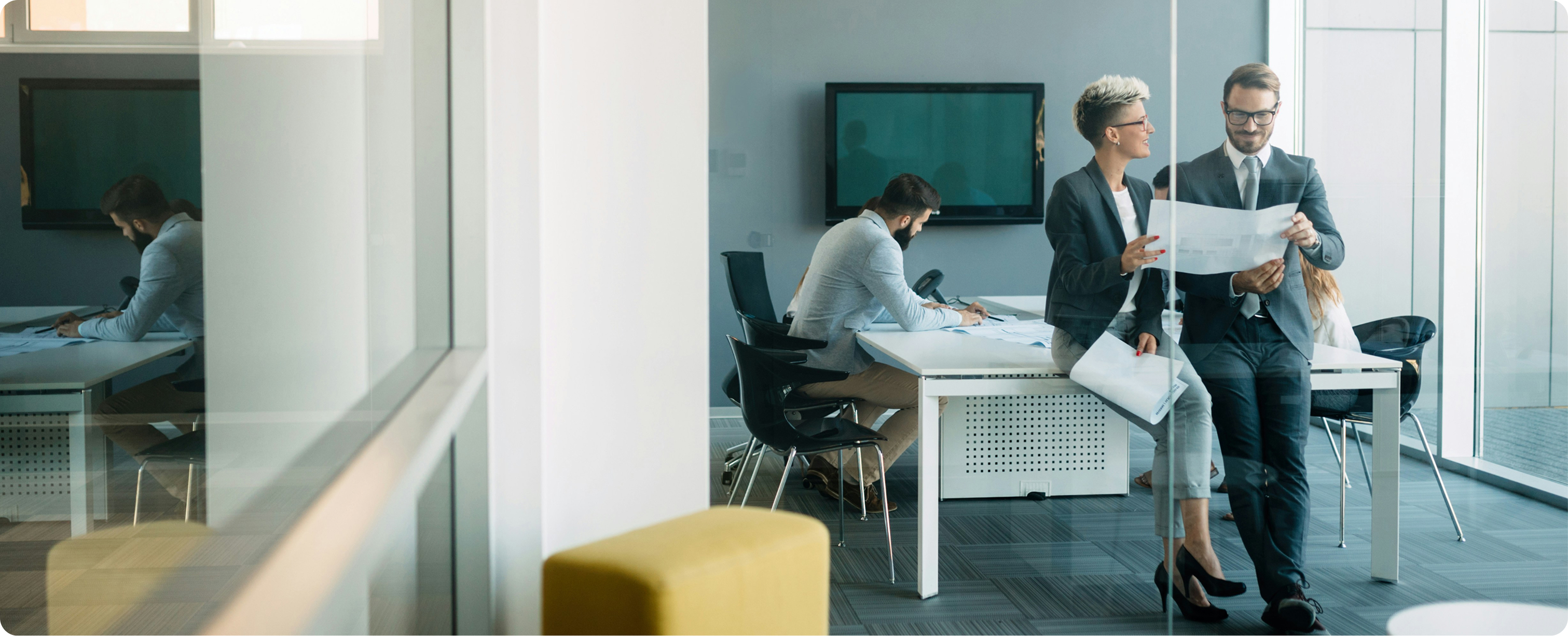 Two professionals, a man and a woman, reviewing documents together near a table while two others work at a desk in a modern office.