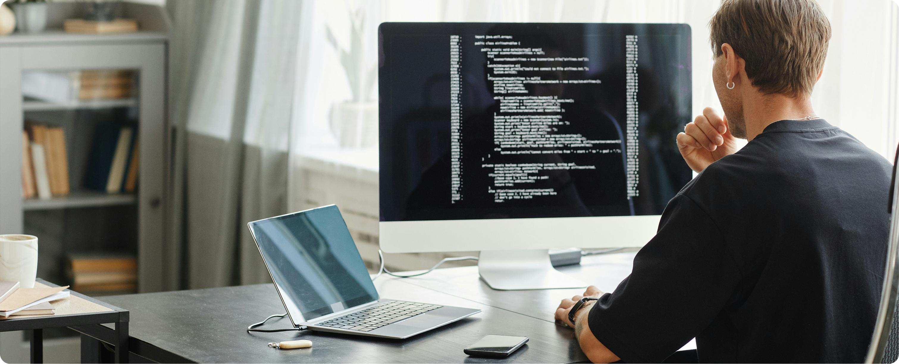 Person sitting at desk looking at a large monitor displaying code, with an open laptop and smartphone on the desk.