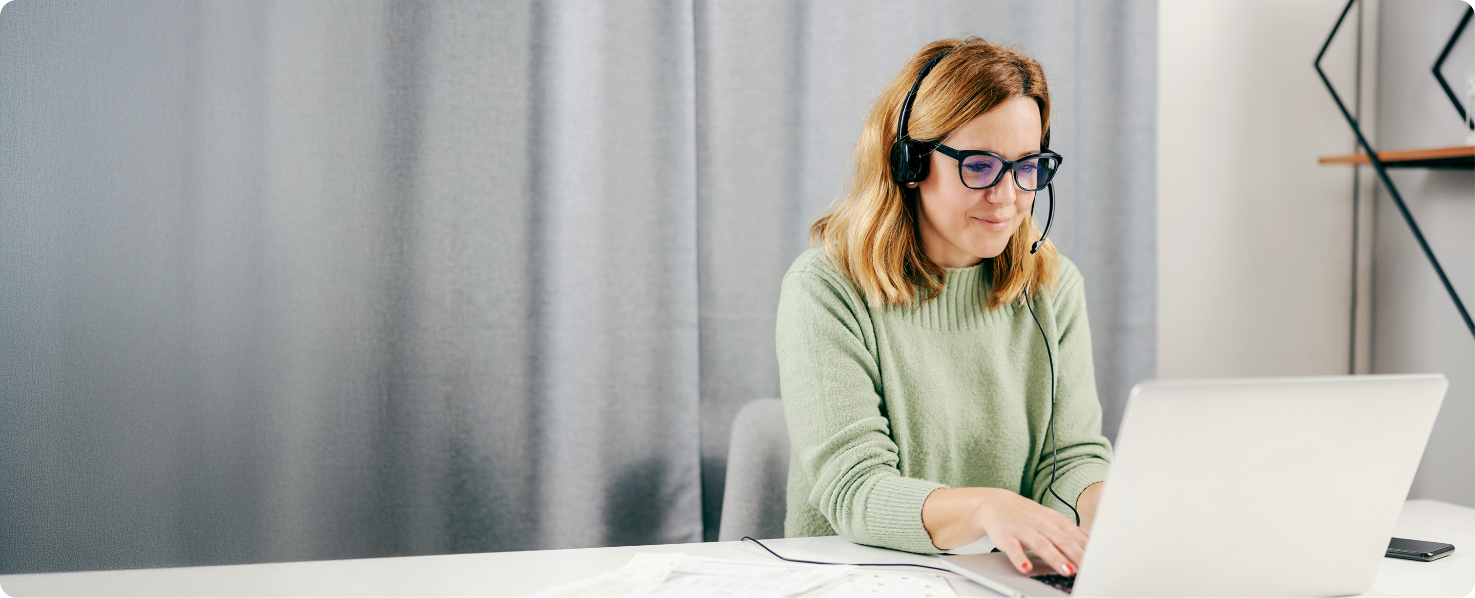 Woman wearing glasses and headset working on a laptop at a desk with documents and smartphone nearby.