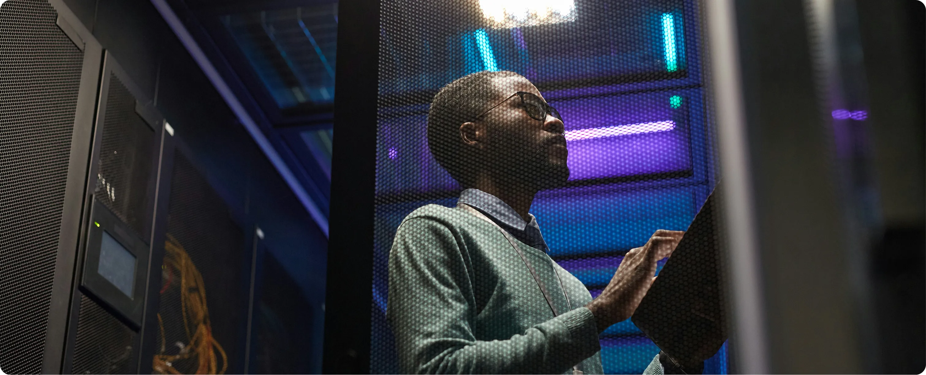 Man wearing glasses working on a laptop inside a server room with blue and purple lights.