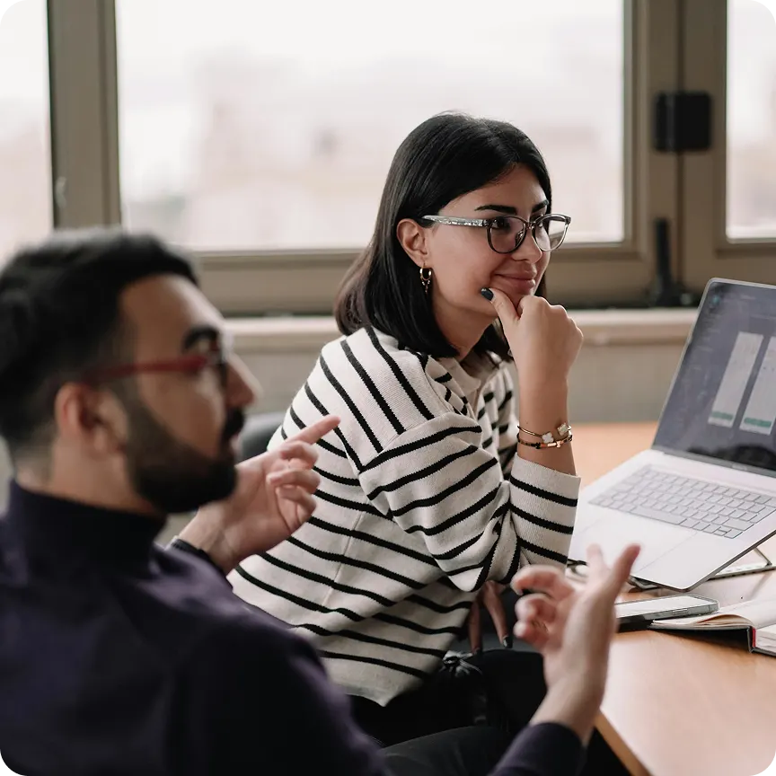 Two colleagues in discussion near a laptop showing charts or graphs in an office setting.