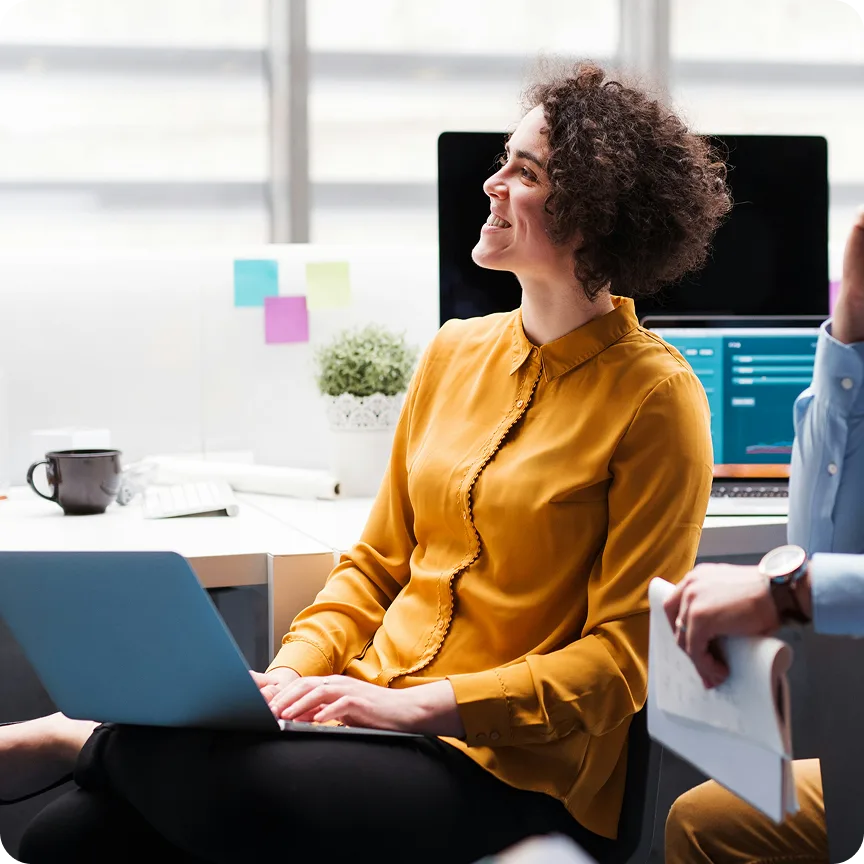 Smiling woman in a mustard yellow blouse working on a laptop in a modern office setting.