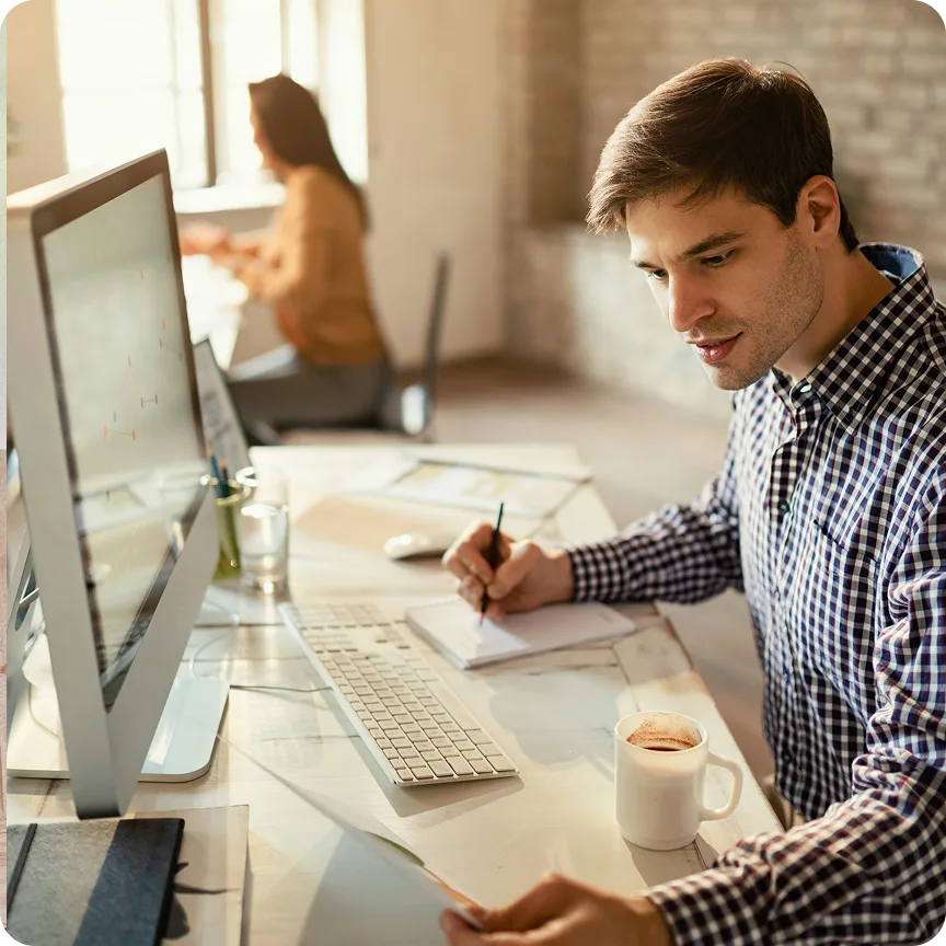 Man in checkered shirt focused on writing notes while working at computer with coffee mug on desk.