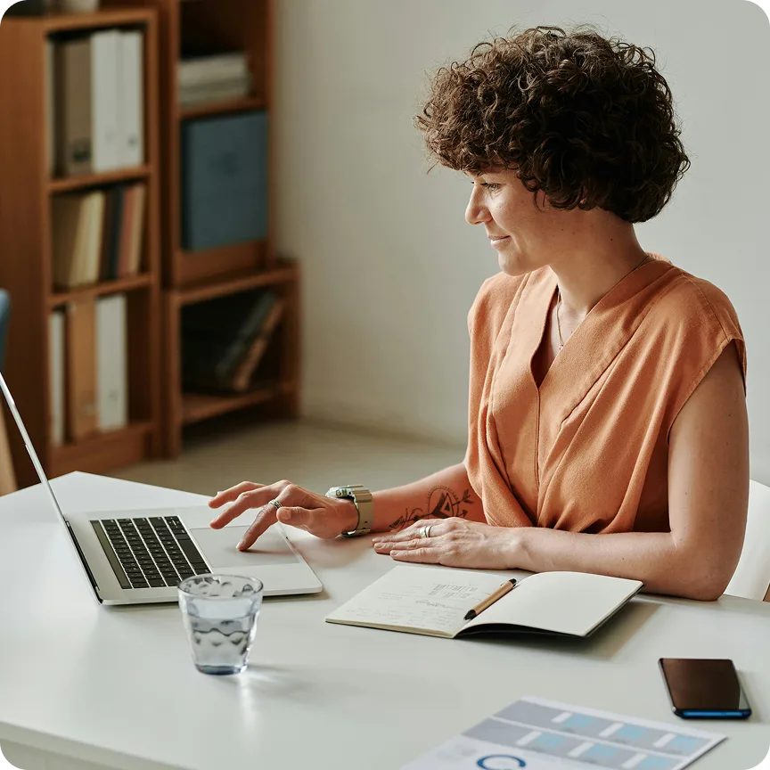 Woman with curly hair sitting at a desk, working on a laptop with a notebook, pen, glass of water, and phone nearby.