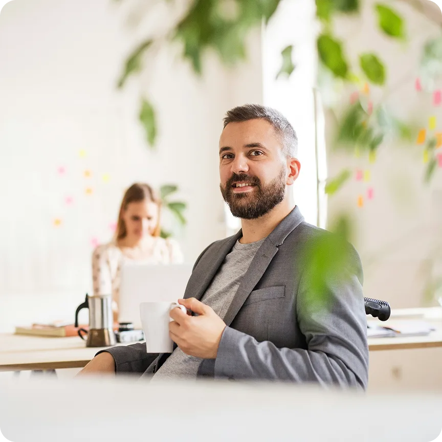 Smiling man in a gray blazer holding a white mug in a bright office with a woman working in the background.