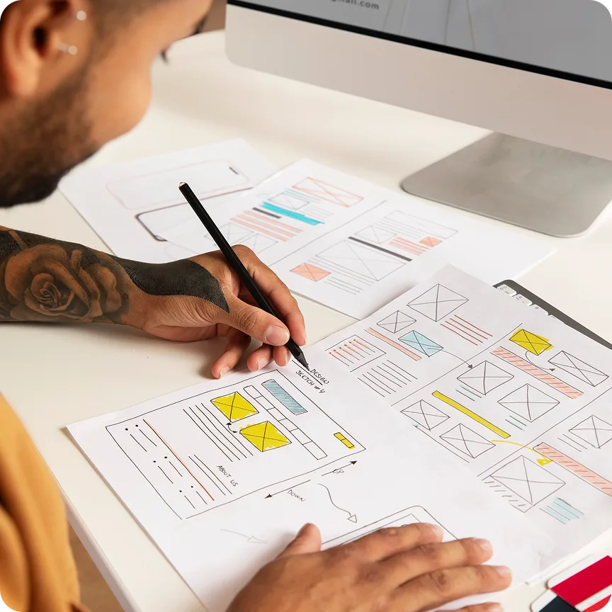 Person with tattooed arm sketching website wireframes on paper at a desk with a computer.