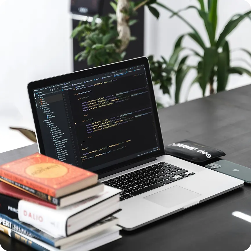 Open laptop displaying code editor on a desk with a stack of books and a smartphone beside it.