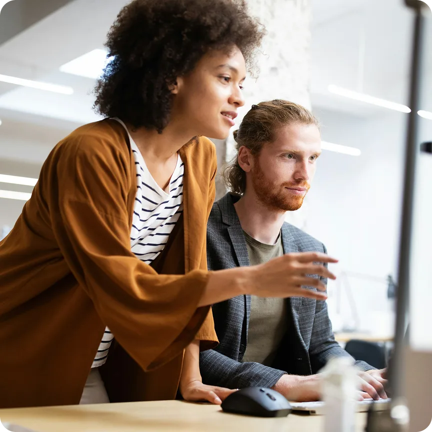 A woman in a brown jacket leans over and points at a computer screen while a man with red hair and beard looks attentively at the screen in a modern office.