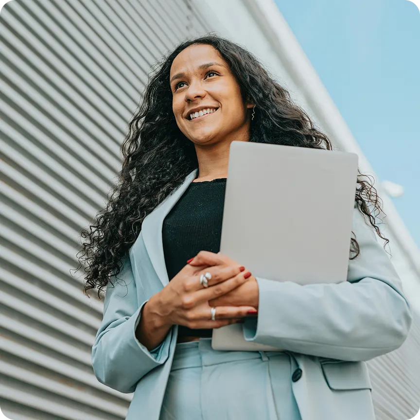 Smiling woman in a light blue suit holding a closed laptop outdoors with a modern building background.