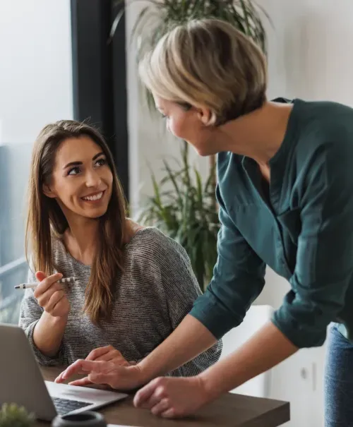 Two women discussing work with laptop open, one sitting and holding pen, other leaning in.
