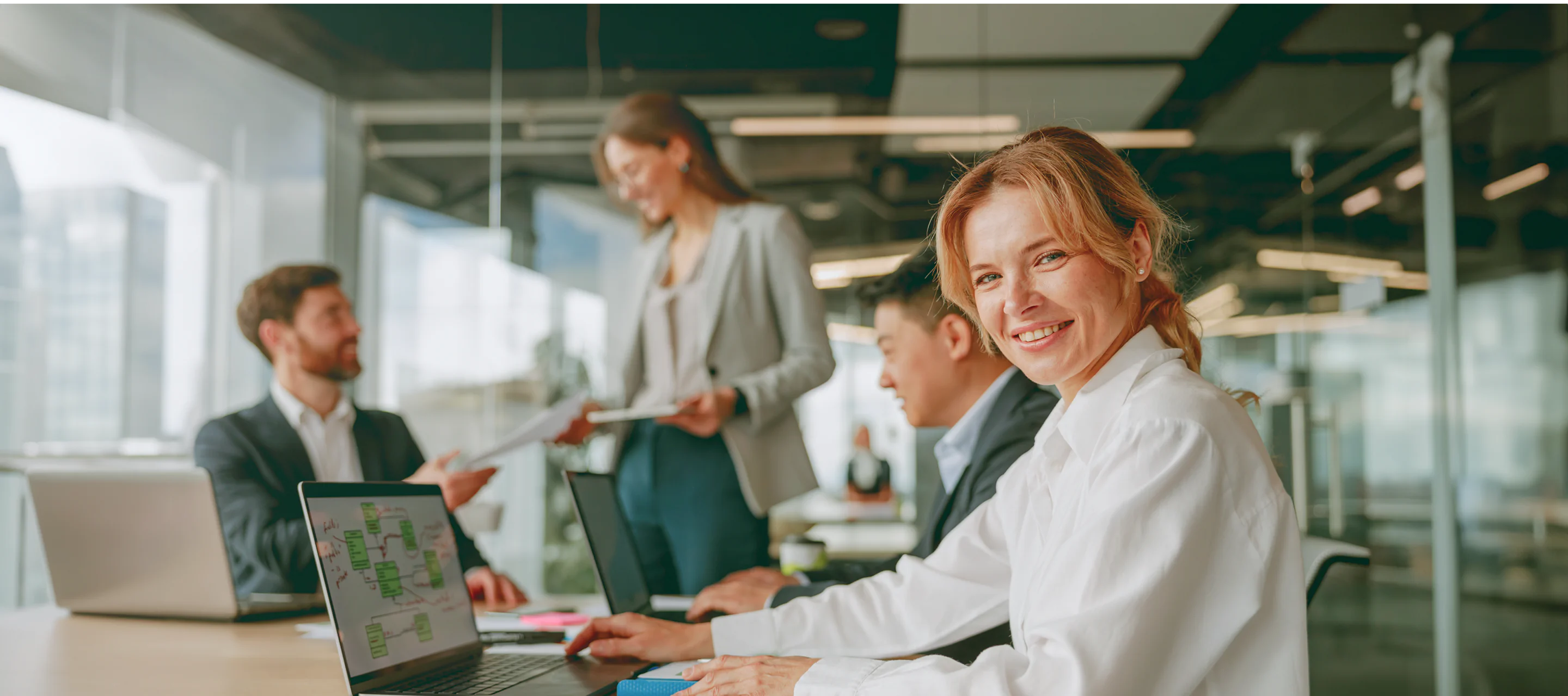 Smiling businesswoman working on a laptop in a modern office with colleagues discussing in the background.