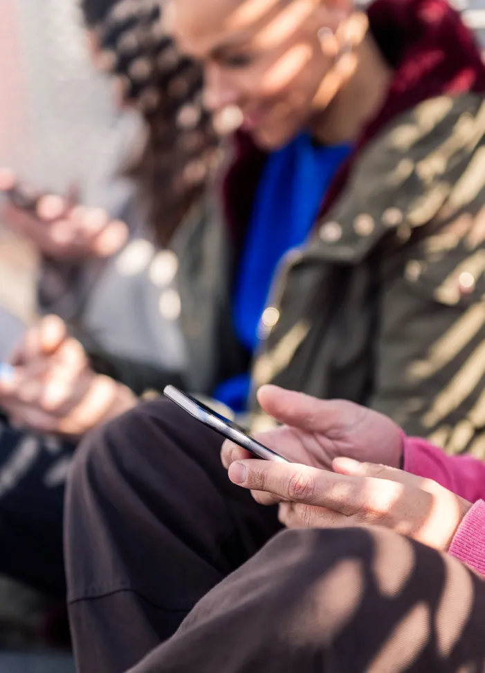 People sitting close together, holding and using smartphones outdoors in dappled sunlight.