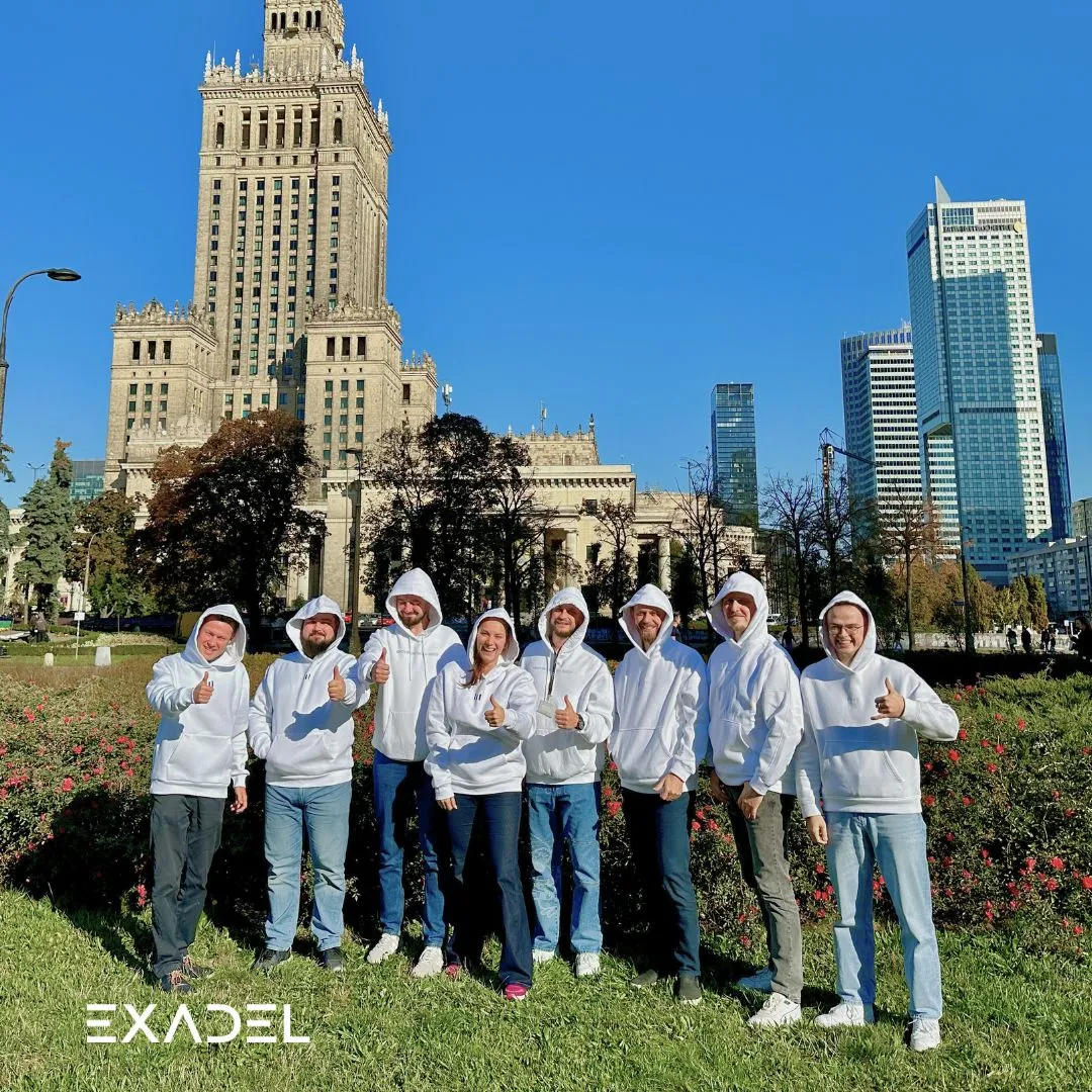 Group of eight people in white hoodies giving thumbs up in a city park with tall buildings behind.