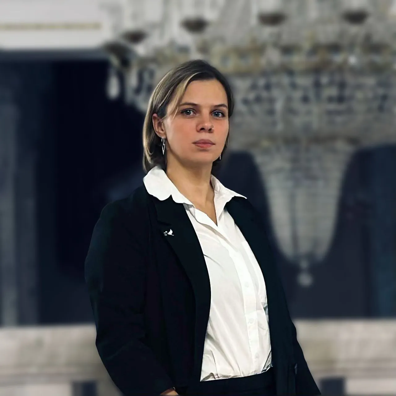 Confident woman in a white shirt and black blazer standing indoors with a blurred chandelier in the background.