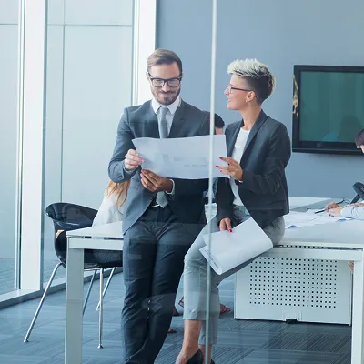 Two business professionals reviewing documents while standing near an office desk with colleagues working behind.