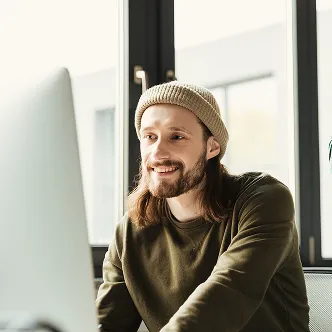 Man with beard and beanie smiling at a computer screen in a modern office.