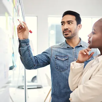 Man in denim shirt writing on glass board while woman in beige jacket watches thoughtfully.