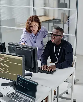 Woman instructing man on coding at a computer desk in a modern office with laptops and monitors.