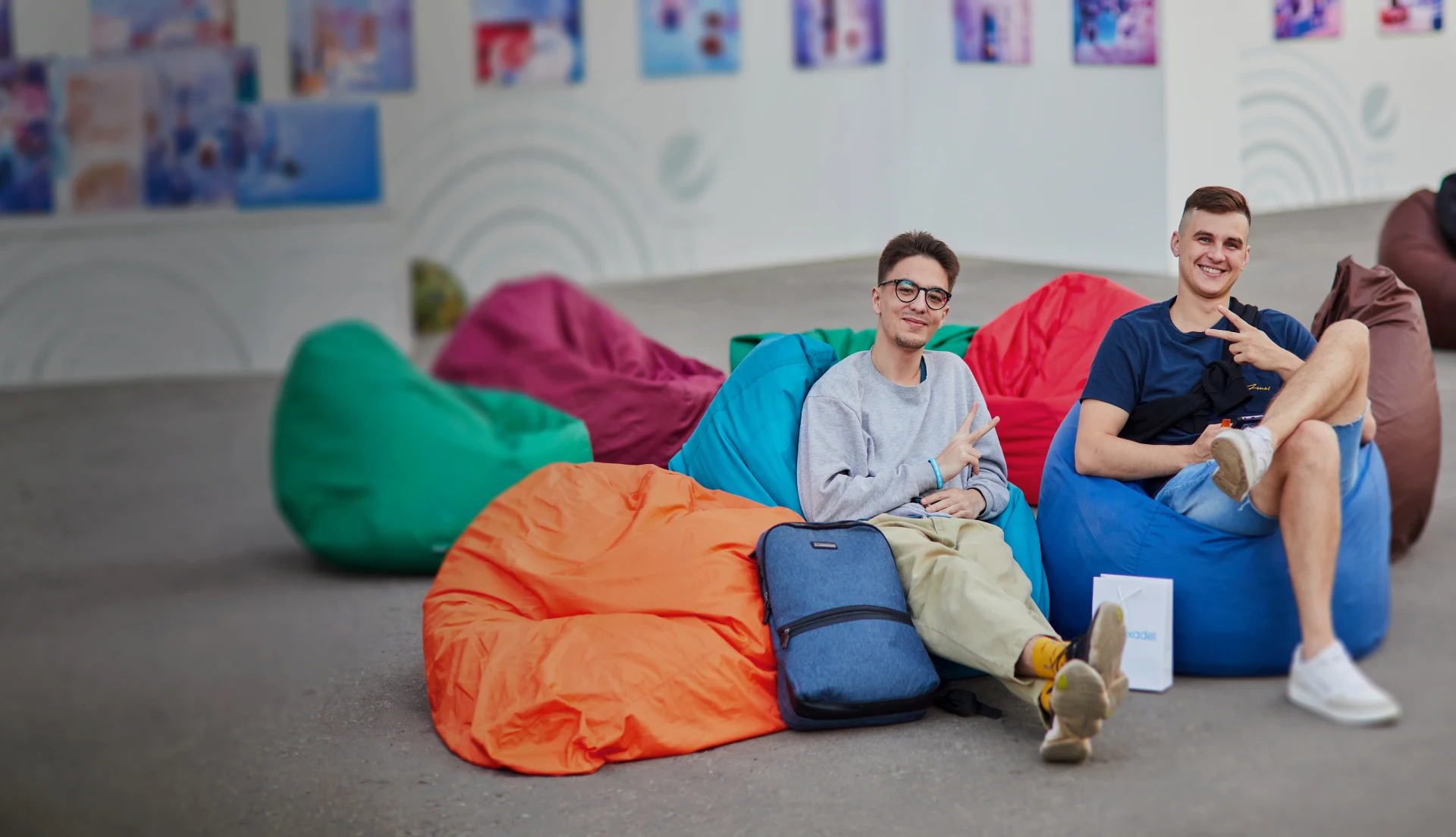Two young men sitting on colorful bean bags, smiling and making peace signs.