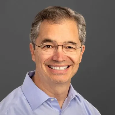 Smiling middle-aged man with salt-and-pepper hair wearing glasses and a light blue shirt.