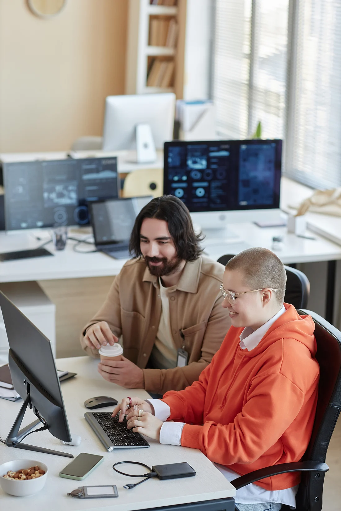 Two colleagues at office desk; one types on keyboard while the other holds a coffee cup.