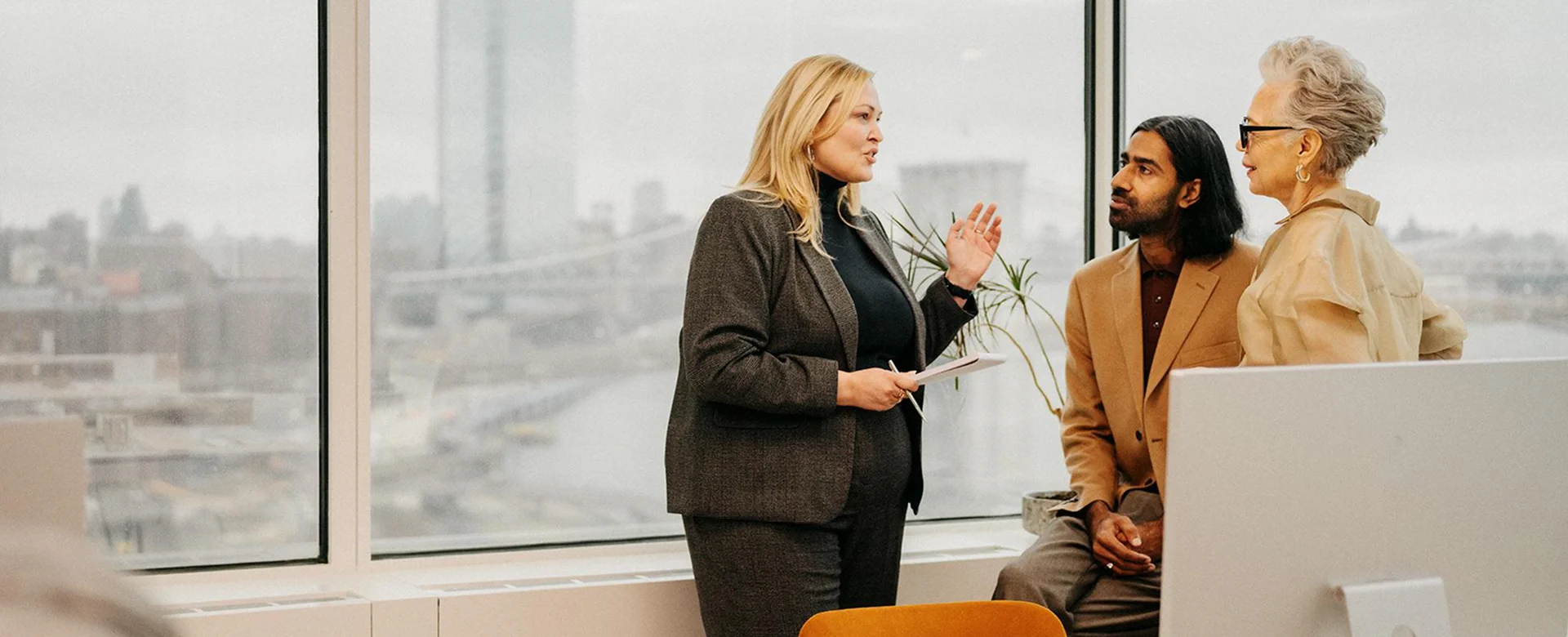 Three professionals standing and talking in an office with large windows overlooking a cityscape.