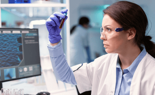 Female scientist in gloves and goggles examining a blood sample in a lab setting with computer screens.