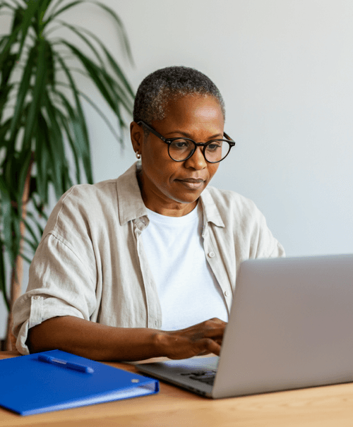 Middle-aged woman with glasses working on a laptop at a wooden desk with a blue folder and pen nearby.