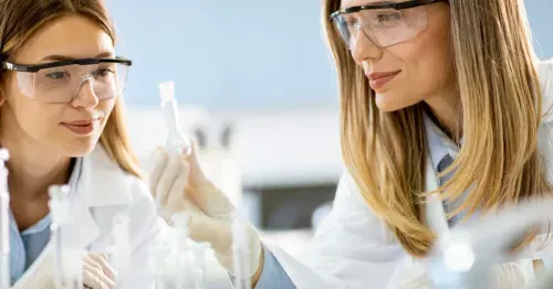 Two female scientists wearing safety goggles examining a flask with liquid in a laboratory.
