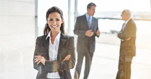 Smiling businesswoman in suit with arms crossed, two men in suits talking blurred in background.