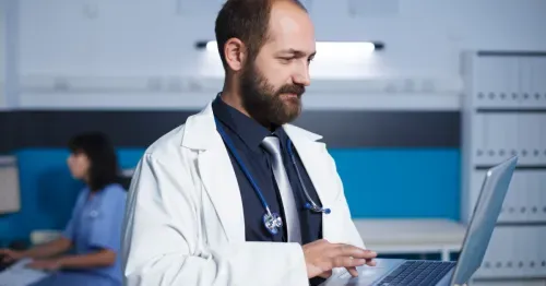 Male doctor in white coat using laptop in a medical office with a nurse in the background.