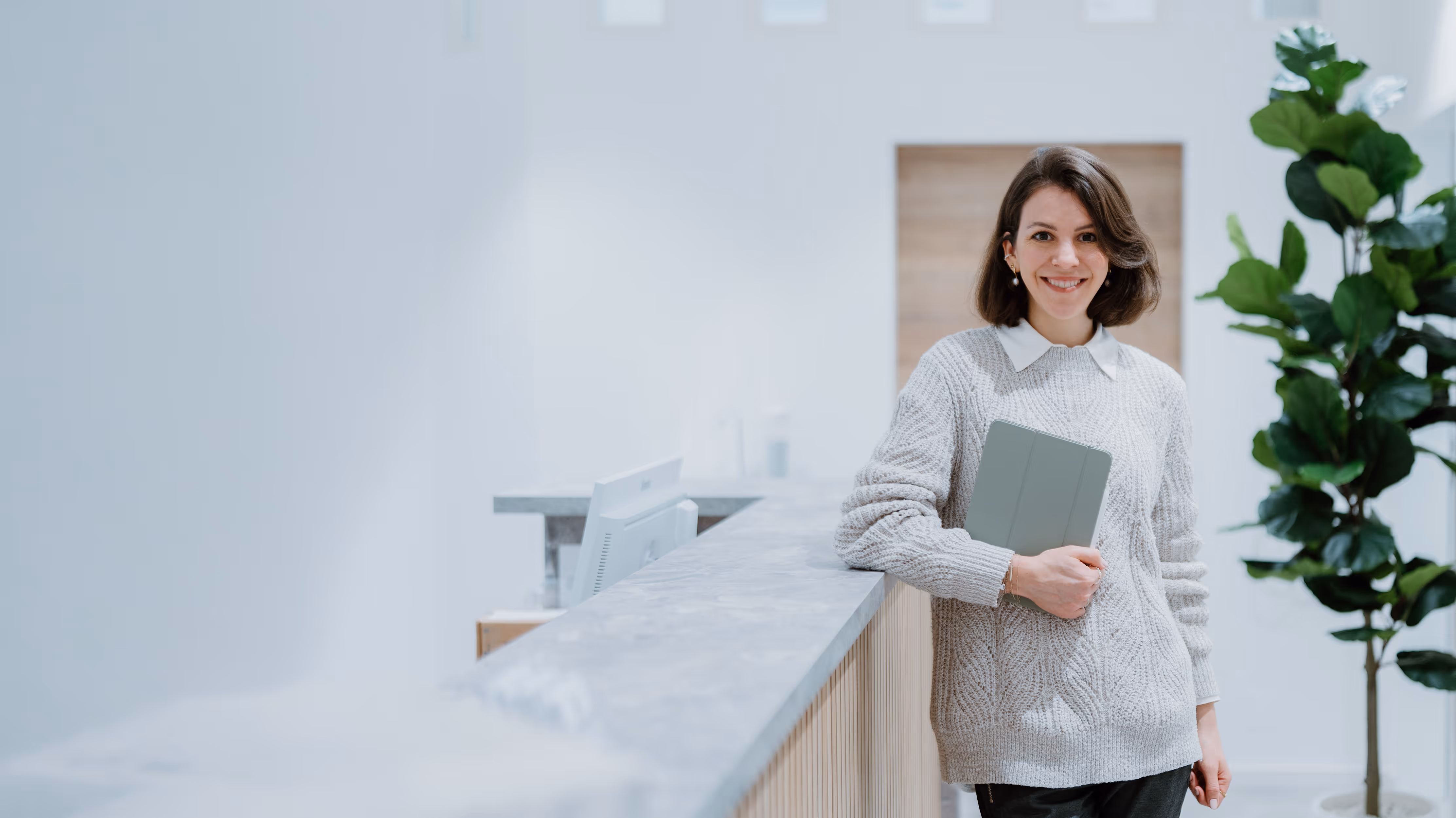 Junge Frau in hellem Pullover lehnt an einer Marmor-Theke und hält ein Tablet, im modernen Büro mit Pflanze im Hintergrund.