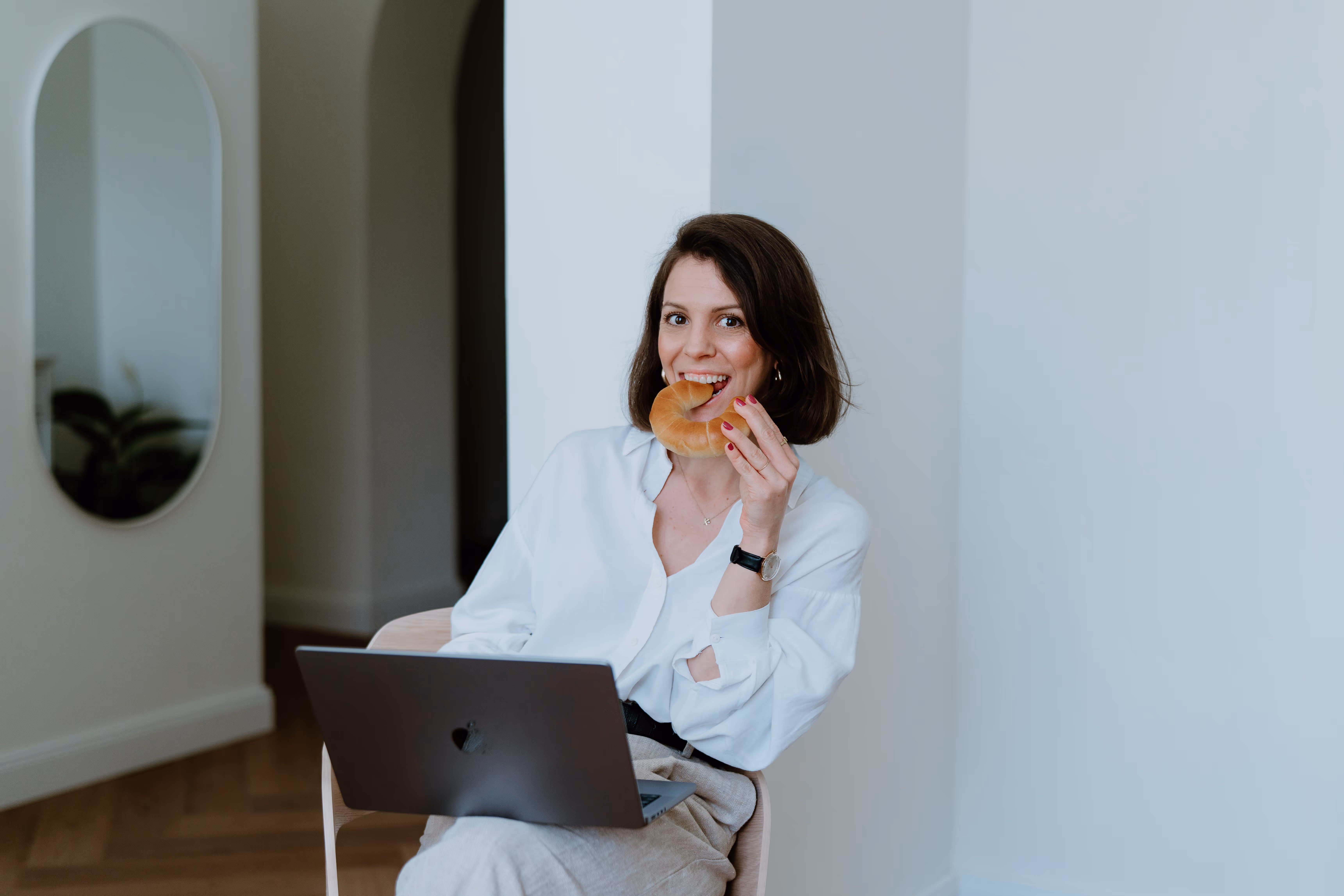 Frau mit weißer Bluse sitzt mit einem Laptop auf dem Schoß auf einem Stuhl und isst einen Croissant.