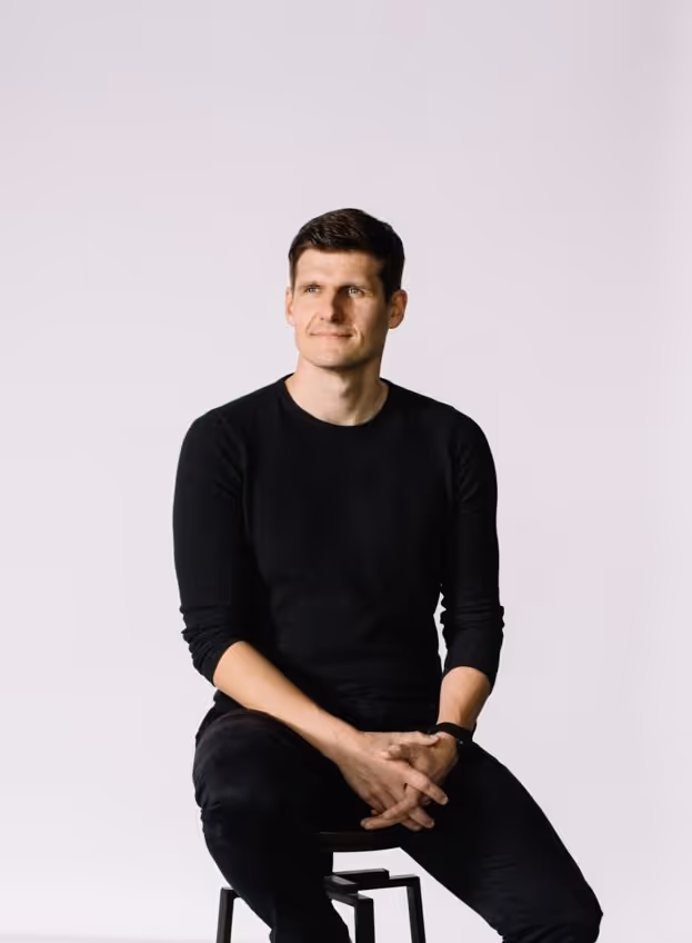 Man with brown hair wearing a black long-sleeve shirt and black pants sitting on a stool against a white background, hands clasped.