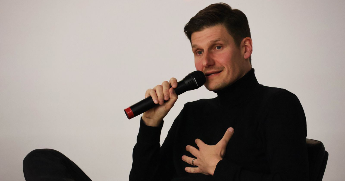 Man in black turtleneck sitting with leg crossed, speaking into a handheld microphone with one hand on his chest.