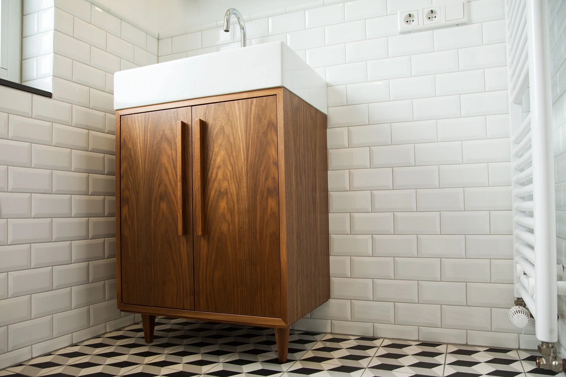 Modernes Badezimmer mit einem weißen Waschbecken auf einem Holzunterschrank, weißen Fliesen an den Wänden und schwarz-weiß gemustertem Boden.