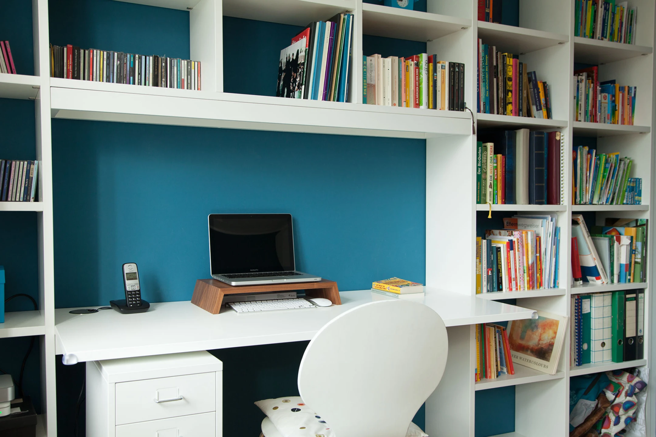 Moderner Arbeitsbereich mit weißem Schreibtisch, Laptop auf Holzständer, Bücherschrank und einem weißen Stuhl mit Kissen.