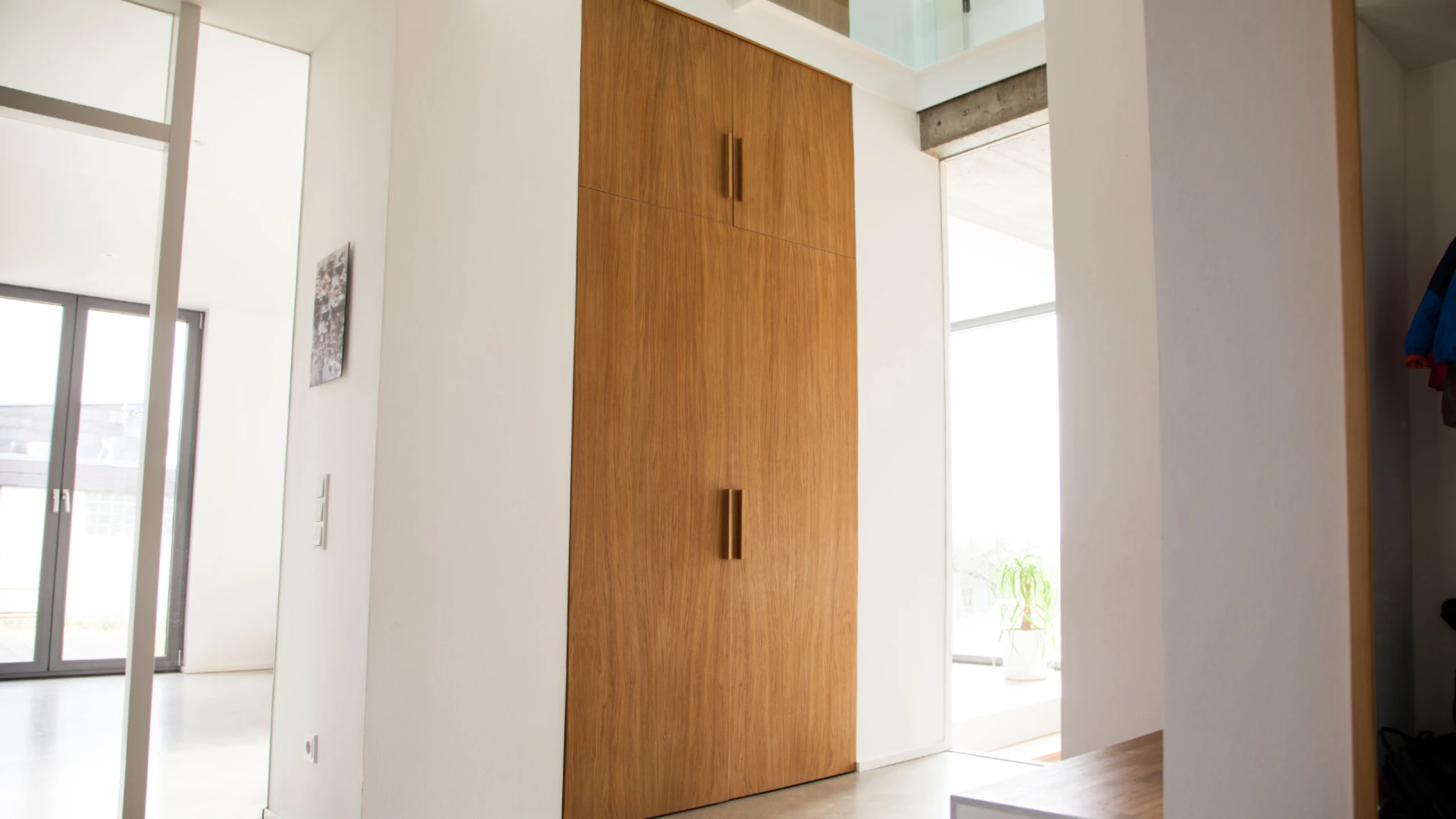 Moderner Innenraum mit einer großen, zweiflügeligen Holztür in einem weißen Flur, daneben Glasfront und Pflanzen.