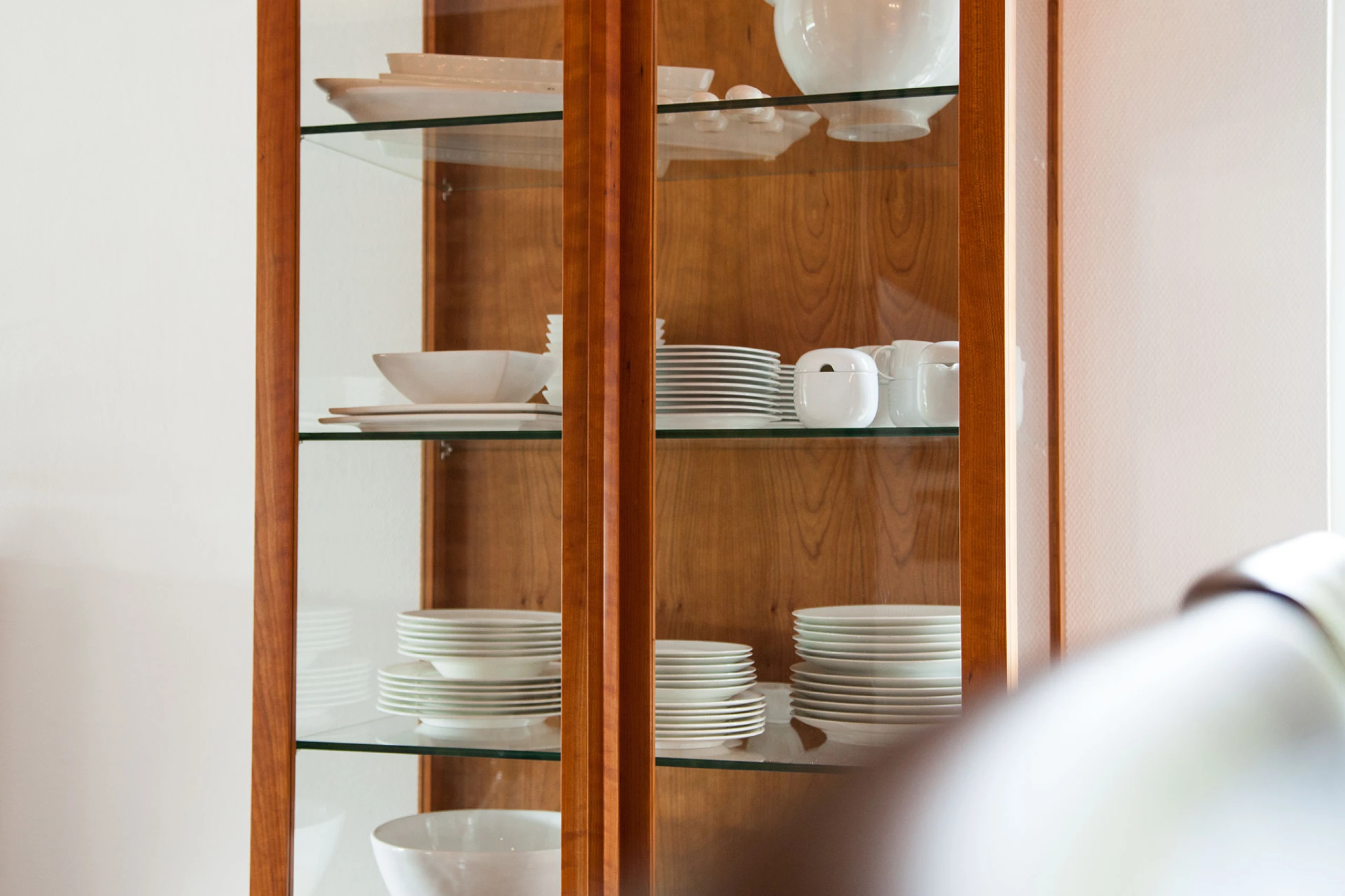 Glasvitrine mit weißen Tellerstapeln, Schalen und Zuckerdosen vor Holzrückwand.