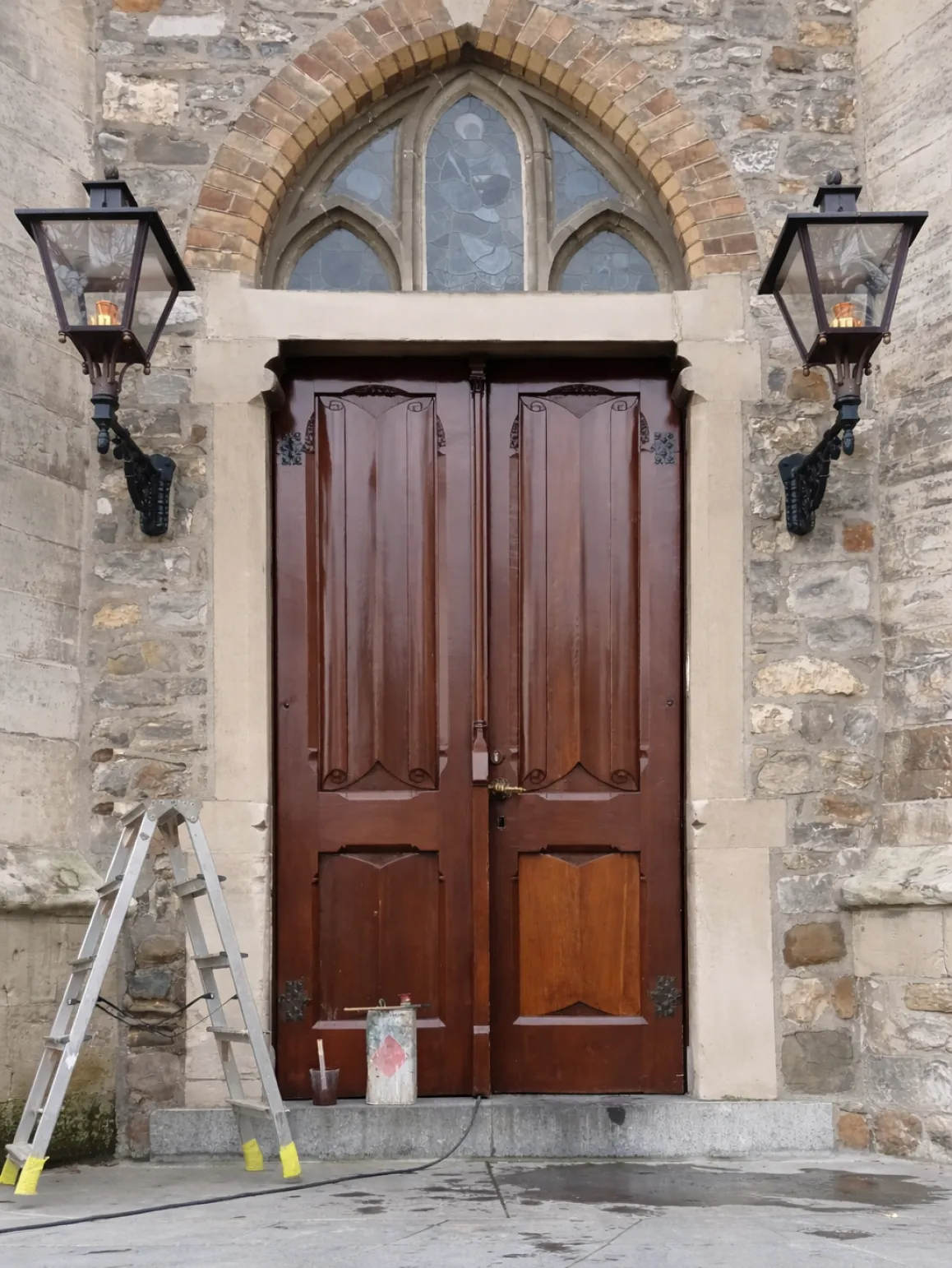 Große braune Holztür mit gotischem Bogenfenster darüber, flankiert von zwei Wandlaternen, eine Leiter und Malutensilien davor.