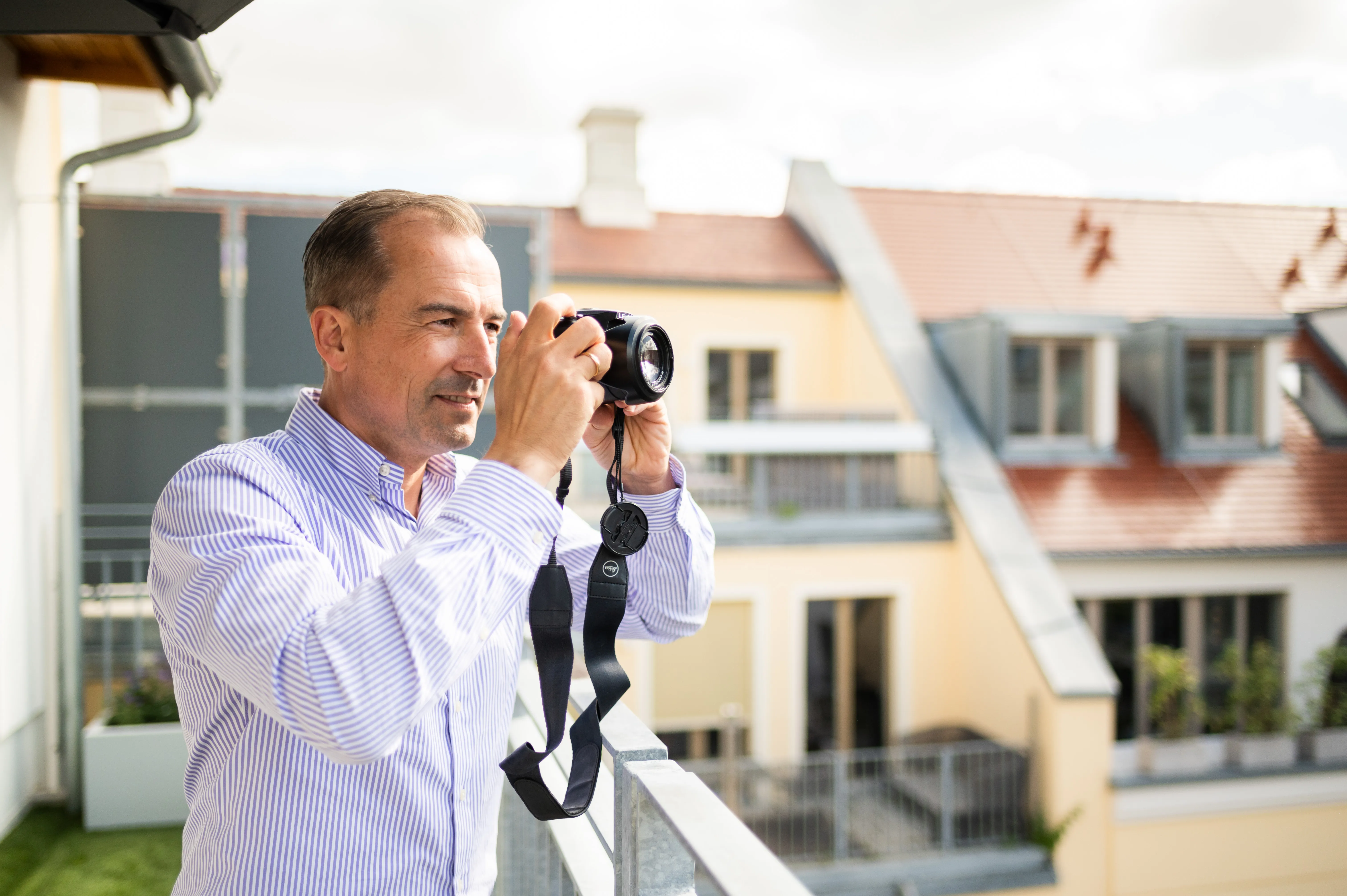 Mann auf Balkon fotografiert mit Kamera, im Hintergrund Wohnhäuser mit Fenstern und Balkon.