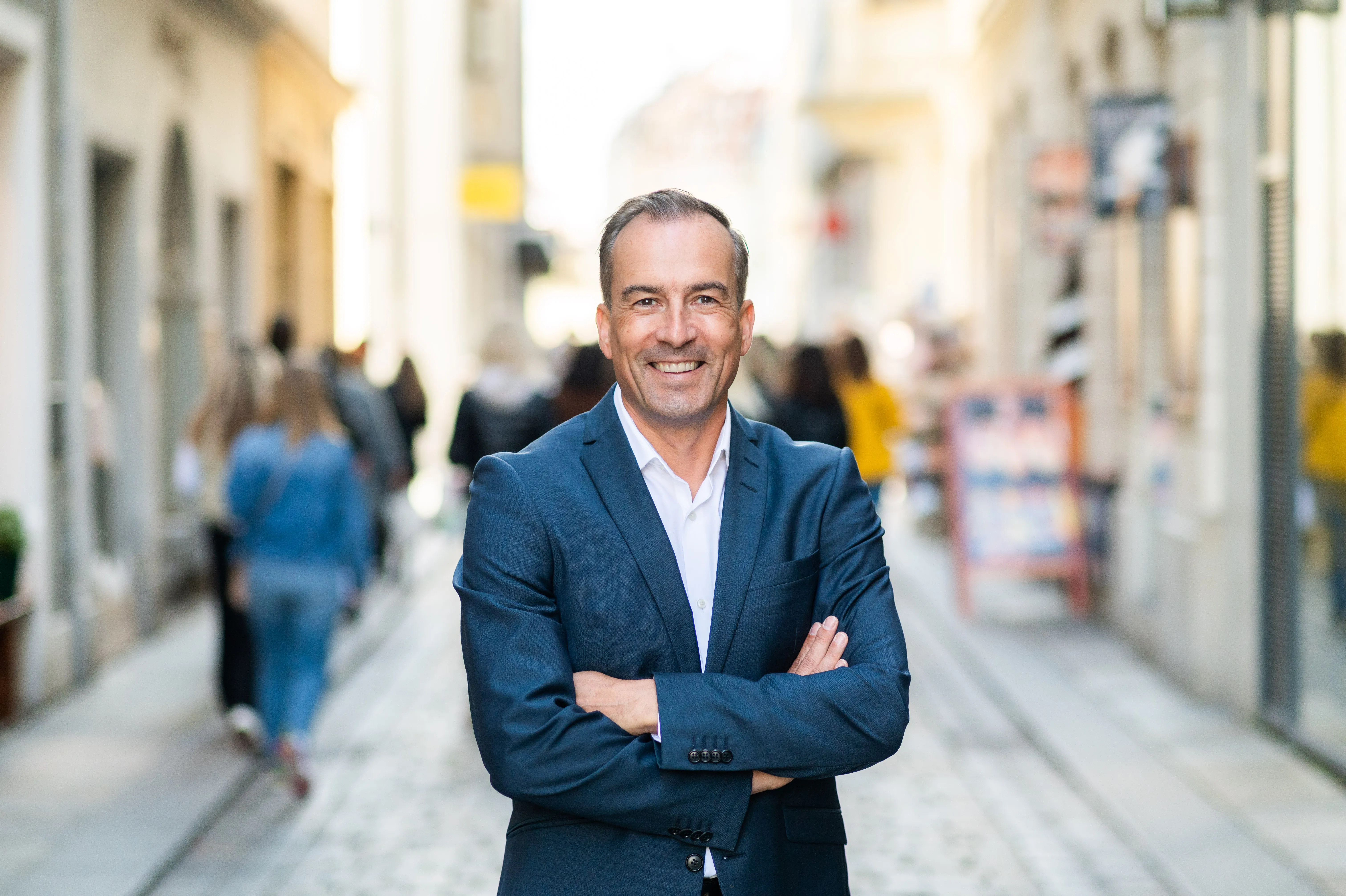 Mann in dunkelblauem Anzug lächelt mit verschränkten Armen auf einer belebten Straße.
