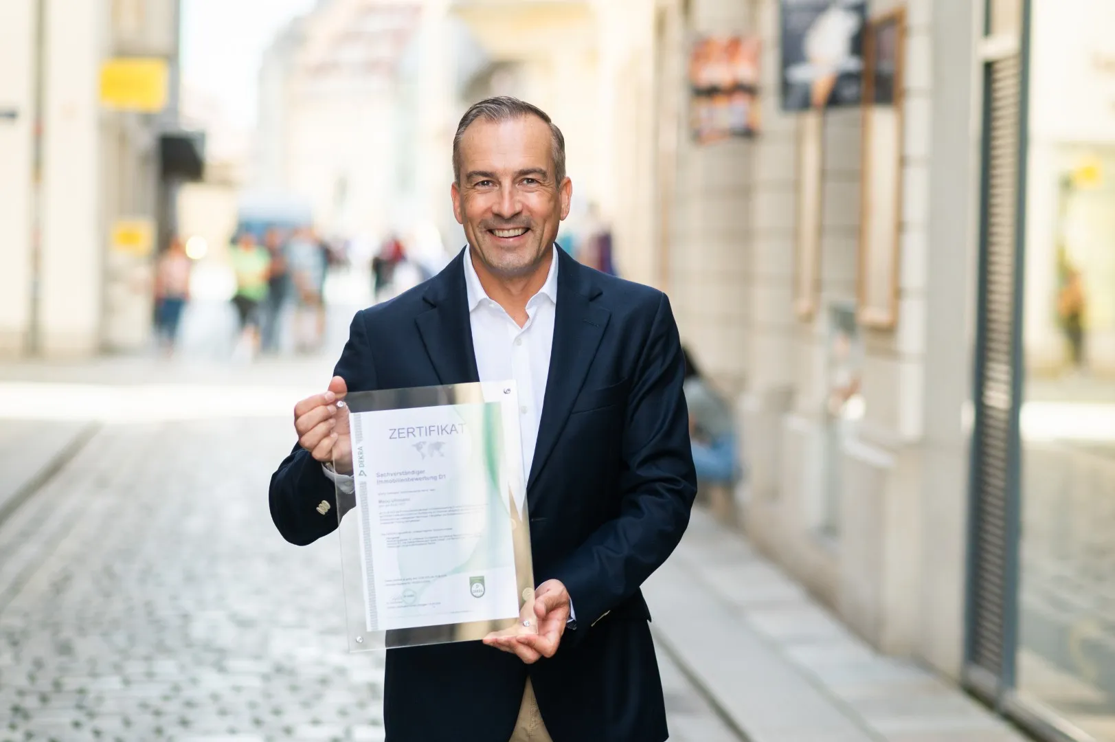 Mann in dunklem Sakko hält lächelnd ein eingerahmtes Zertifikat in einer belebten Fußgängerzone.