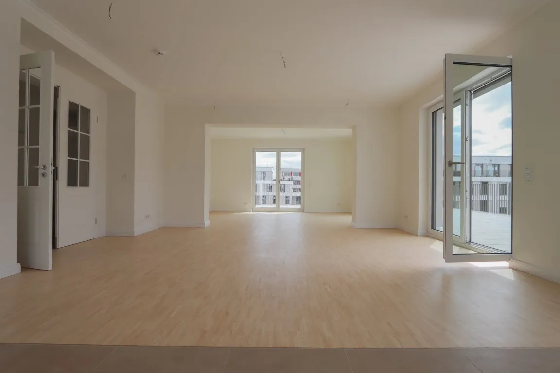 Leerer, lichtdurchfluteter Raum mit hellem Holzfußboden, weißen Wänden und großen Fenstern mit Blick auf Nachbargebäude.
