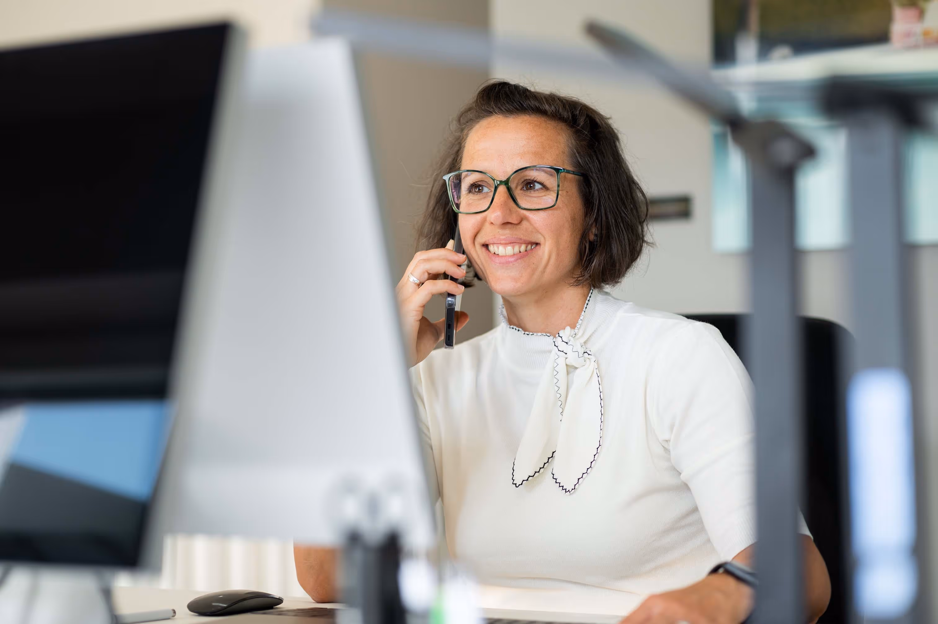 Lächelnde Frau mit Brille telefoniert am Schreibtisch vor Computerbildschirm.
