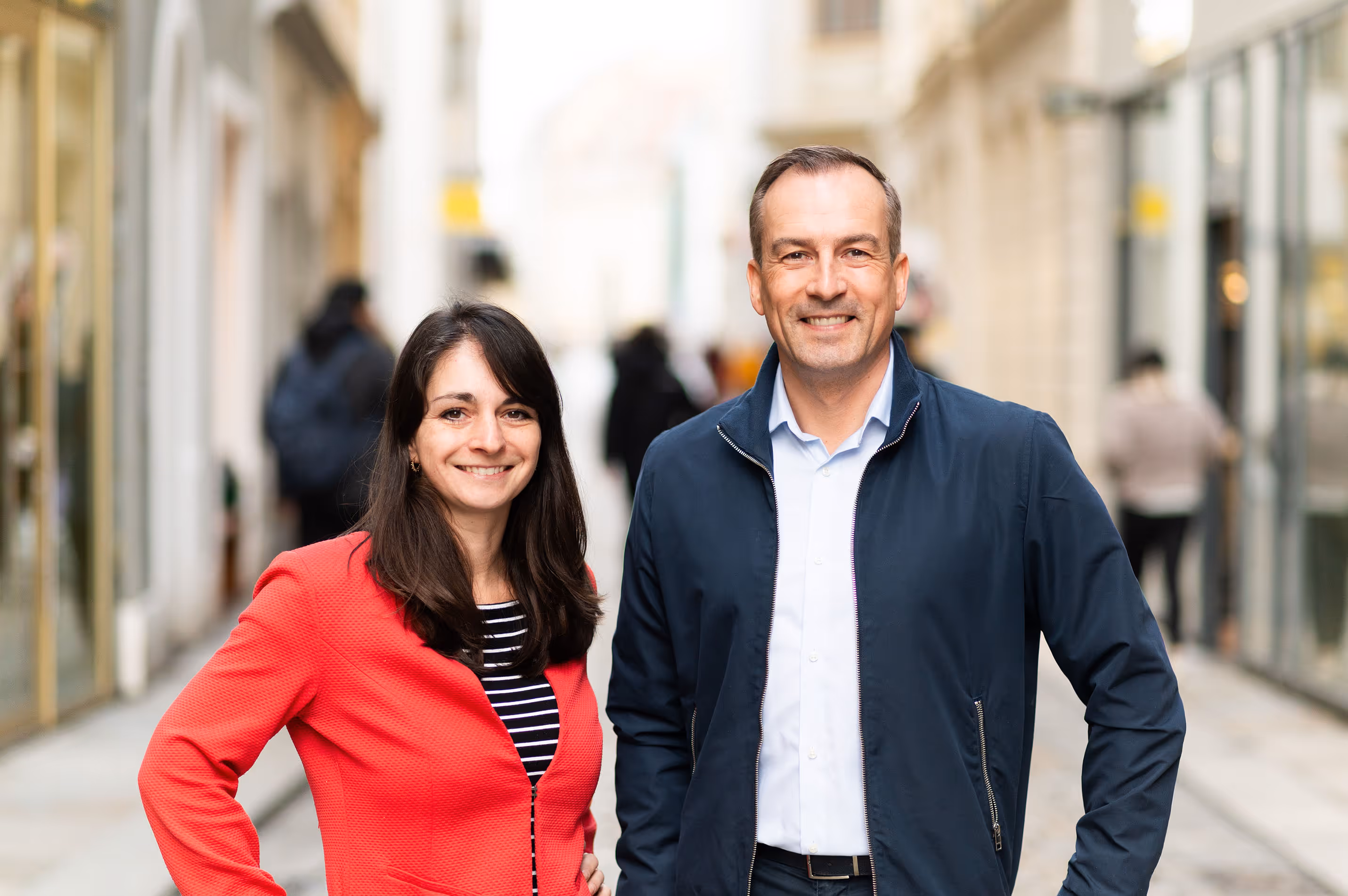 Zwei lächelnde Personen, eine Frau in rotem Blazer und ein Mann in dunkelblauer Jacke, stehen nebeneinander in einer Fußgängerzone.