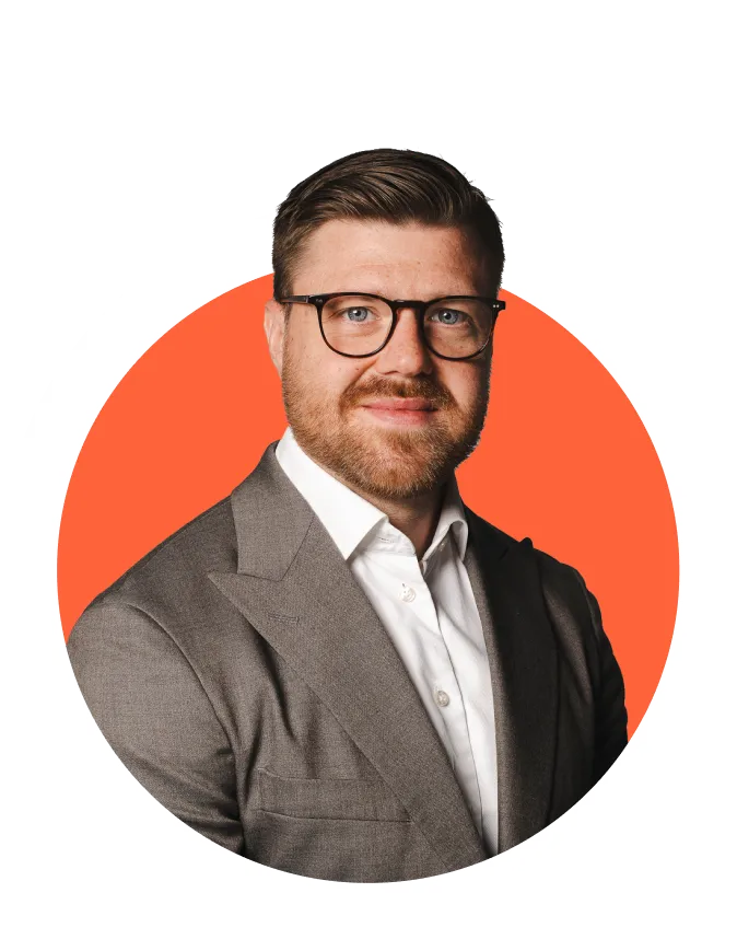 Man with short brown hair, beard, and glasses wearing a gray suit jacket and white shirt, smiling with an orange circular background.