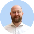 Smiling bald man with a beard wearing a white collared shirt, on a light blue background.